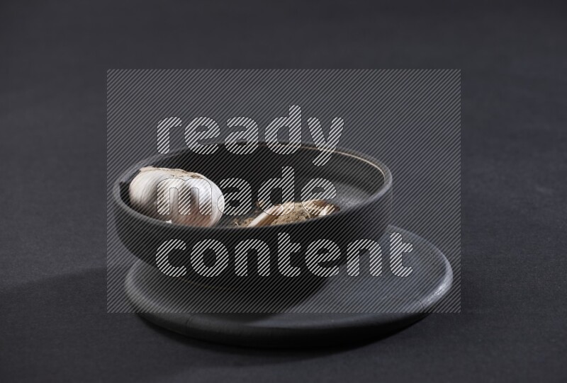 A black pottery bowl full of garlic powder, some cloves and a whole garlic bulb on a black flooring