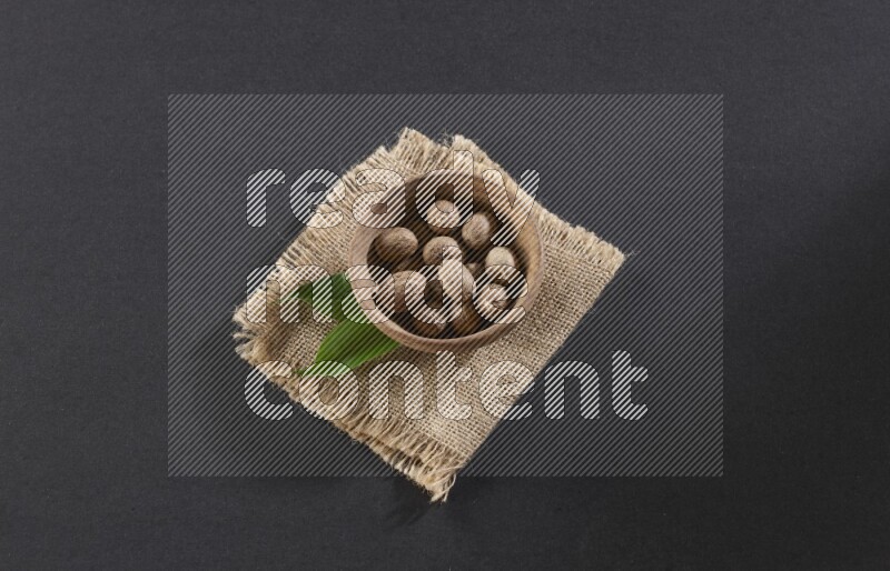 A wooden bowl full of whole nutmeg seeds on burlap fabric on a black flooring