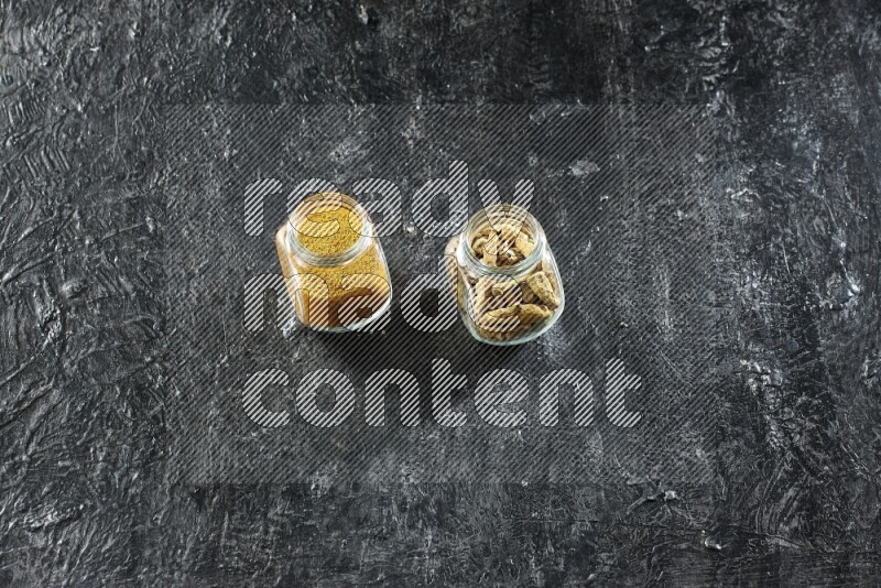 وعاءان زجاجيان للتوابل ممتلئان ببودرة الكركم وأصابع الكركم المجففة على خلفية سوداء