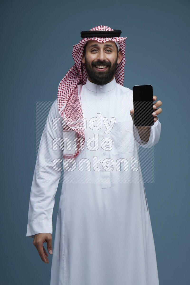 A man showing his phone Screen in a blue background wearing Saudi Thob with Shomag