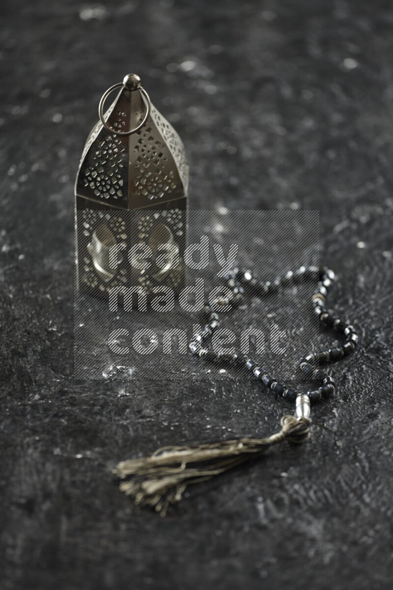 A silver lantern with different drinks, dates, nuts, prayer beads and quran on textured black background