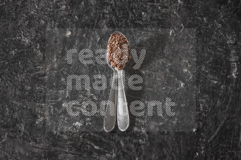 2 metal spoons full of flaxseeds on a textured black flooring
