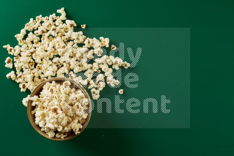 An off white ceramic bowl full of popcorn with popcorn beside it on a green background in a top view shot