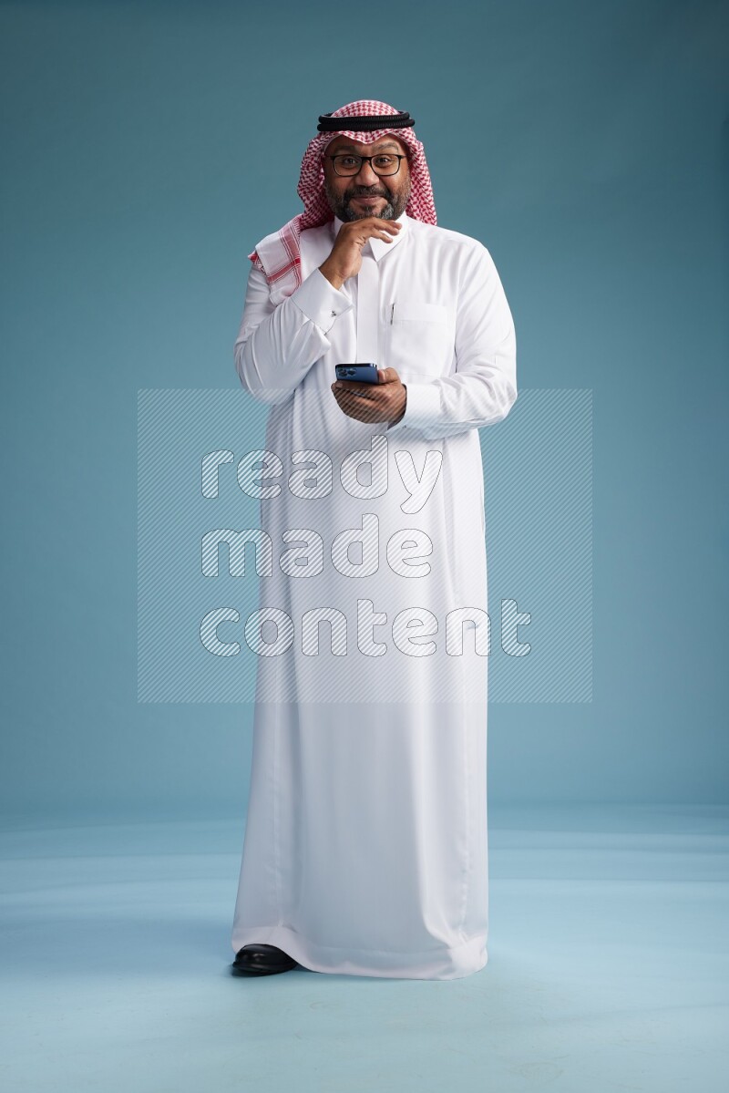 Saudi Man with shimag Standing texting on phone on blue background