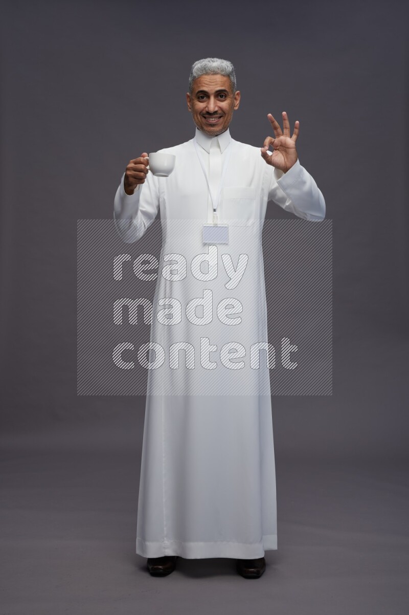 Saudi man wearing thob with neck strap employee badge standing holding mug on gray background
