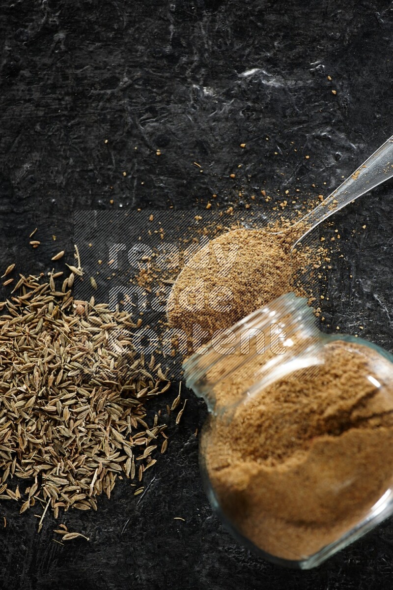 A flipped glass spice jar and a metal spoon full of cumin powder and powder spilled out with cumin seeds on a textured black flooring