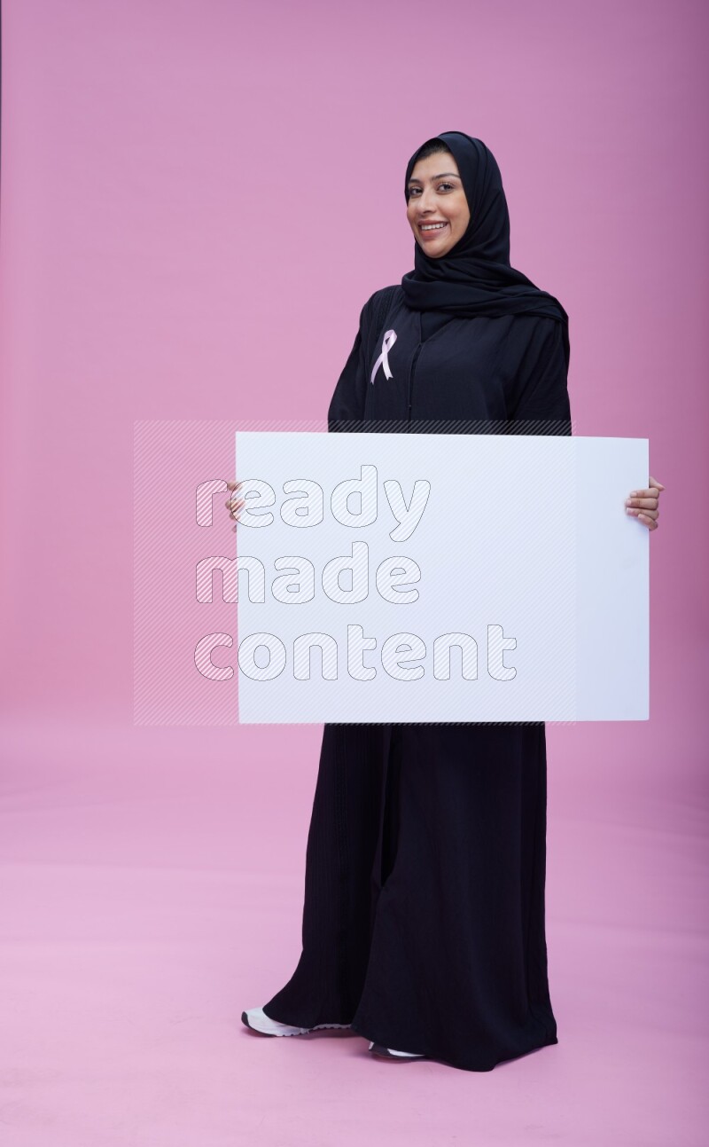 Saudi woman wearing pink ribbon on Abaya standing holding board on pink background