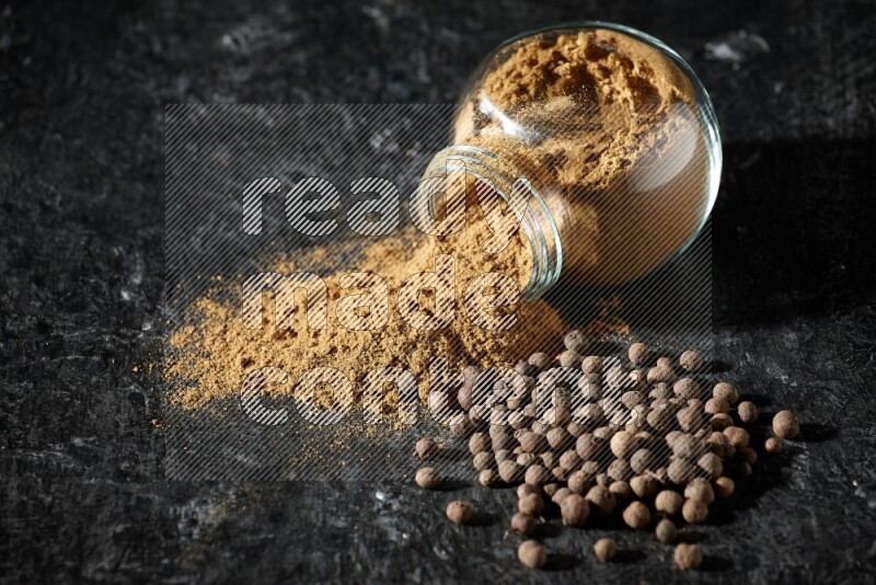 A flipped glass spice jar full of allspice powder and powder spilled out of it with whole balls on a textured black flooring