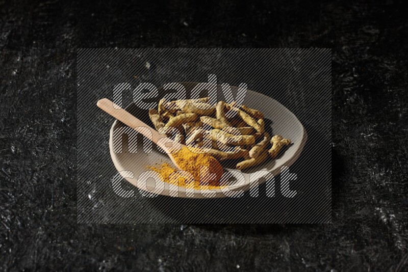 A plate filled with dried turmeric fingers and a wooden spoon full of turmeric powder on a textured black flooring
