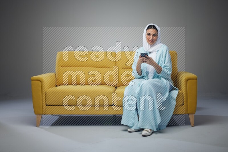 A Saudi woman wearing a light blue Abaya and white head scarf sitting on a yellow sofa and using her phone eye level on a grey background
