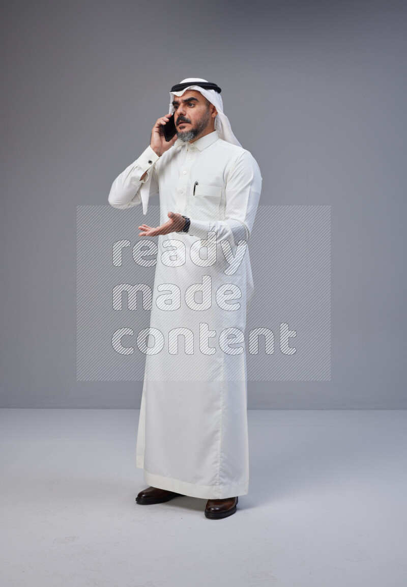Saudi man Wearing Thob and white Shomag standing talking on phone on Gray background