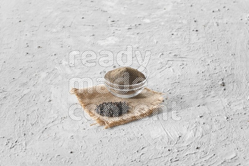 A glass bowl full of black pepper powder and black pepper beads on burlap fabric on textured white flooring