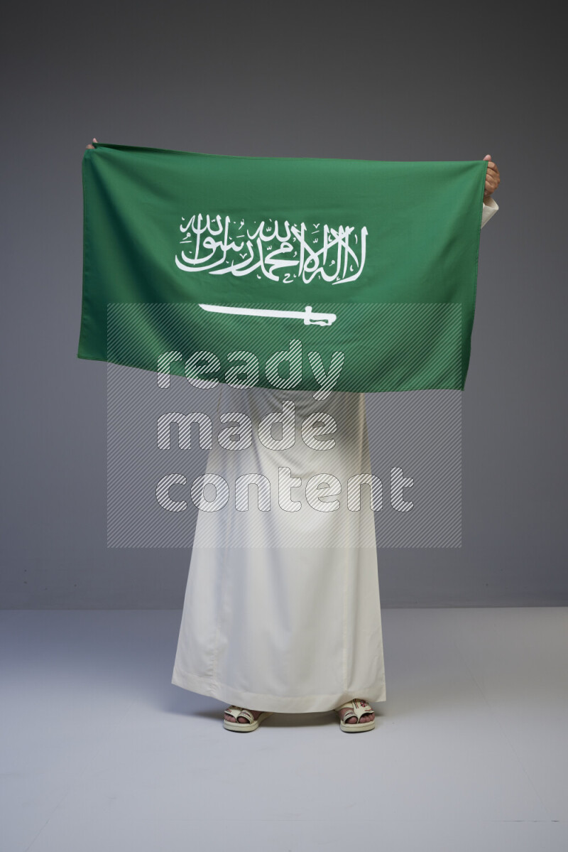 A Saudi man standing wearing thob holding big Saudi flag on gray background