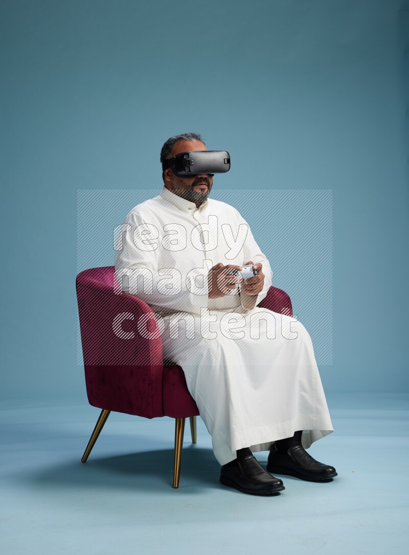 Saudi Man without shimag sitting on chair  gaming with VR and joystick on blue background