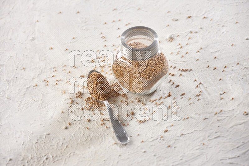 A glass spice jar and a metal spoon full of mustard seeds on a textured white flooring