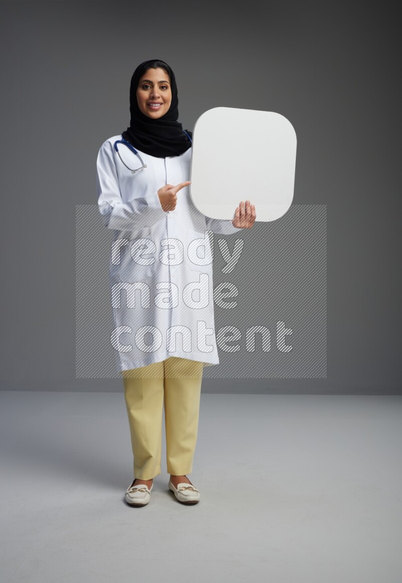 Saudi woman wearing lab coat with stethoscope standing holding social media sign on Gray background