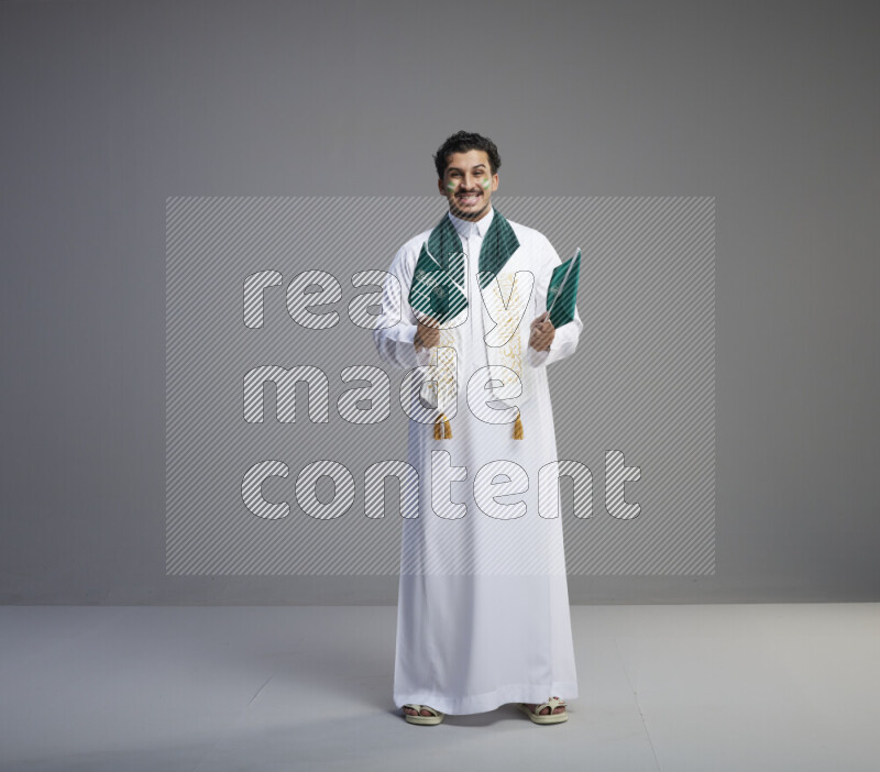 A Saudi man standing wearing thob and saudi flag scarf with face painting holding small Saudi flag on gray background