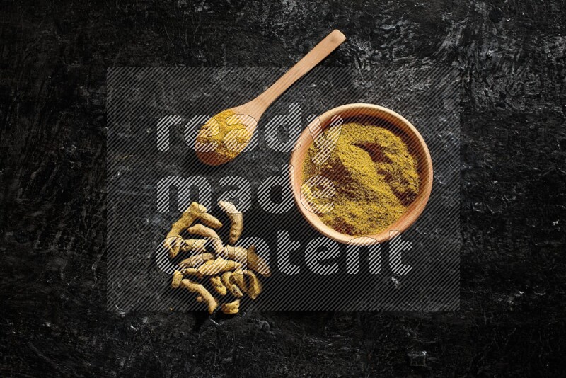 A wooden bowl and a wooden spoon full of turmeric powder with dried turmeric fingers on textured black flooring
