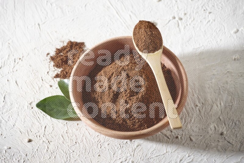 A wooden bowl and a wooden spoon full of cloves powder on a textured white flooring