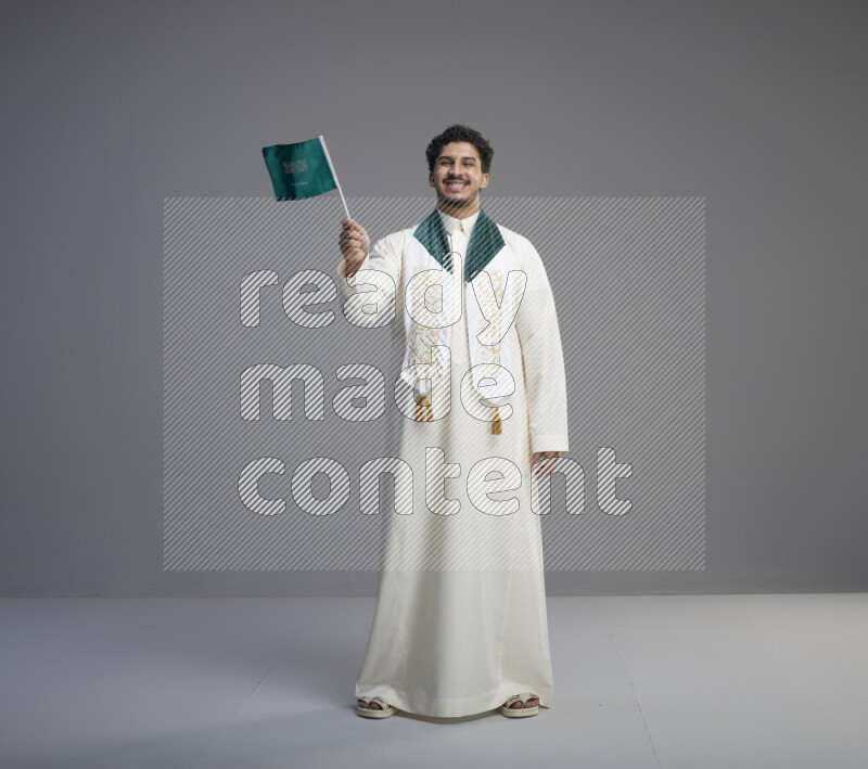 A Saudi man standing wearing thob and saudi flag scarf and holding small saudi flag on gray background