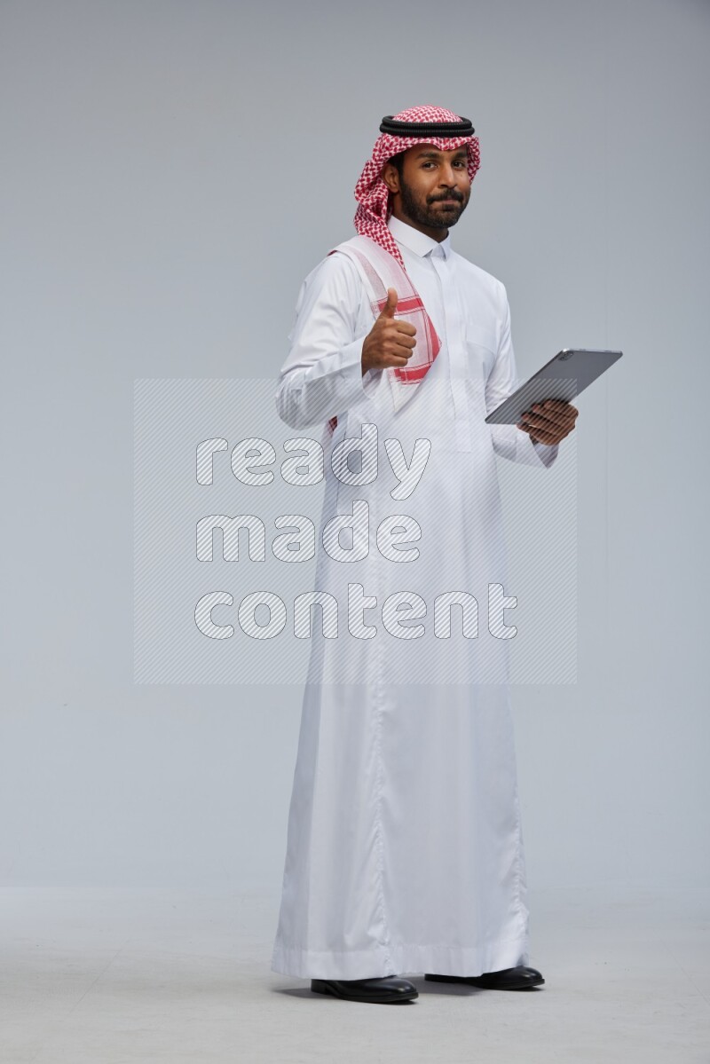 Saudi man Wearing Thob and shomag standing working on tablet on Gray background