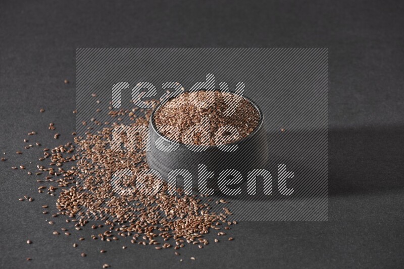 A black pottery bowl full of flaxseeds surrounded by the seeds on a black flooring