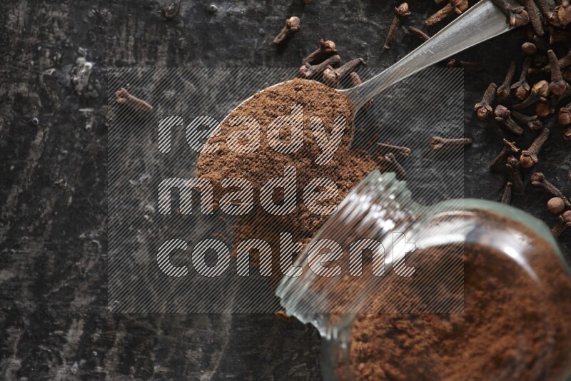 A flipped glass spice jar and a metal spoon full of cloves powder with cloves spread on textured black flooring