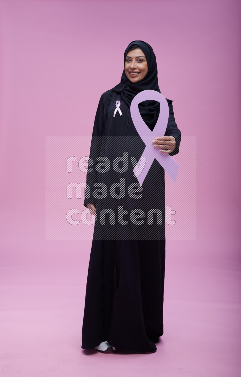 Saudi woman wearing pink ribbon on abaya standing holding awareness ribbon on plain pink background