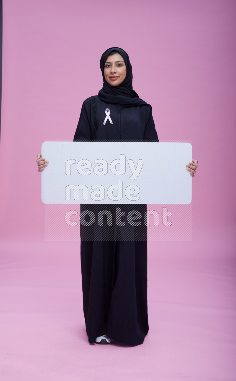 Saudi woman wearing pink ribbon on Abaya standing holding board on pink background