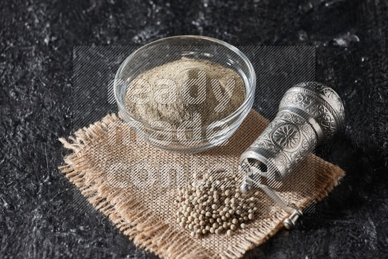 A glass bowl full of white pepper powder with white pepper beads on a burlap piece of fabric and a metal grinder on textured black flooring
