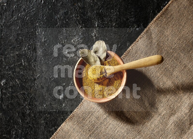A wooden bowl and a wooden spoon full of turmeric powder on burlap fabric on textured black flooring