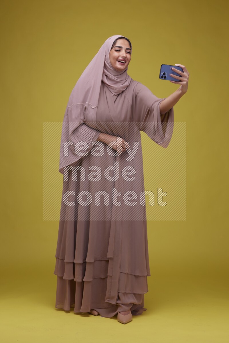 A woman taking a Selfie on a Yellow Background wearing Brown Abaya with Hijab