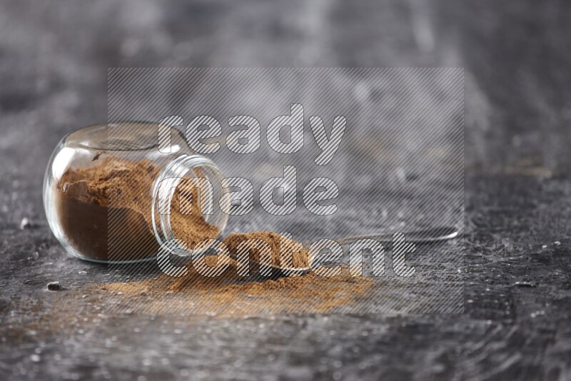 Herbal glass jar full of cinnamon powder flipped and a metal spoon on textured black background