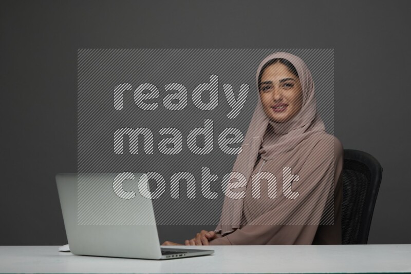 A Saudi woman Setting on her desk on a Gray Background wearing Brown Abaya with Hijab