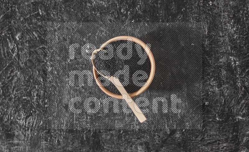 A wooden bowl full of black seeds with wooden spoon full of the seeds on it on a textured black flooring