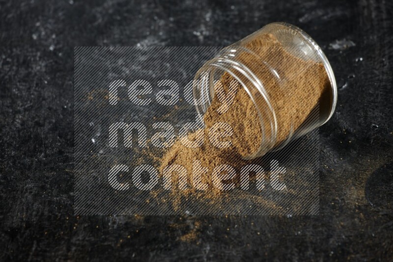 A flipped glass jar full of cumin powder and powder fell out on a textured black flooring