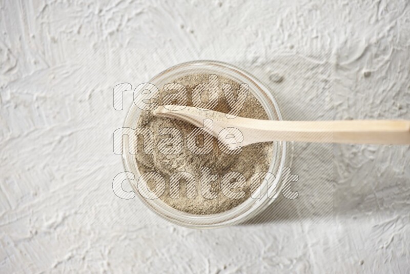 A glass jar and wooden spoon full of white pepper powder on textured white flooring