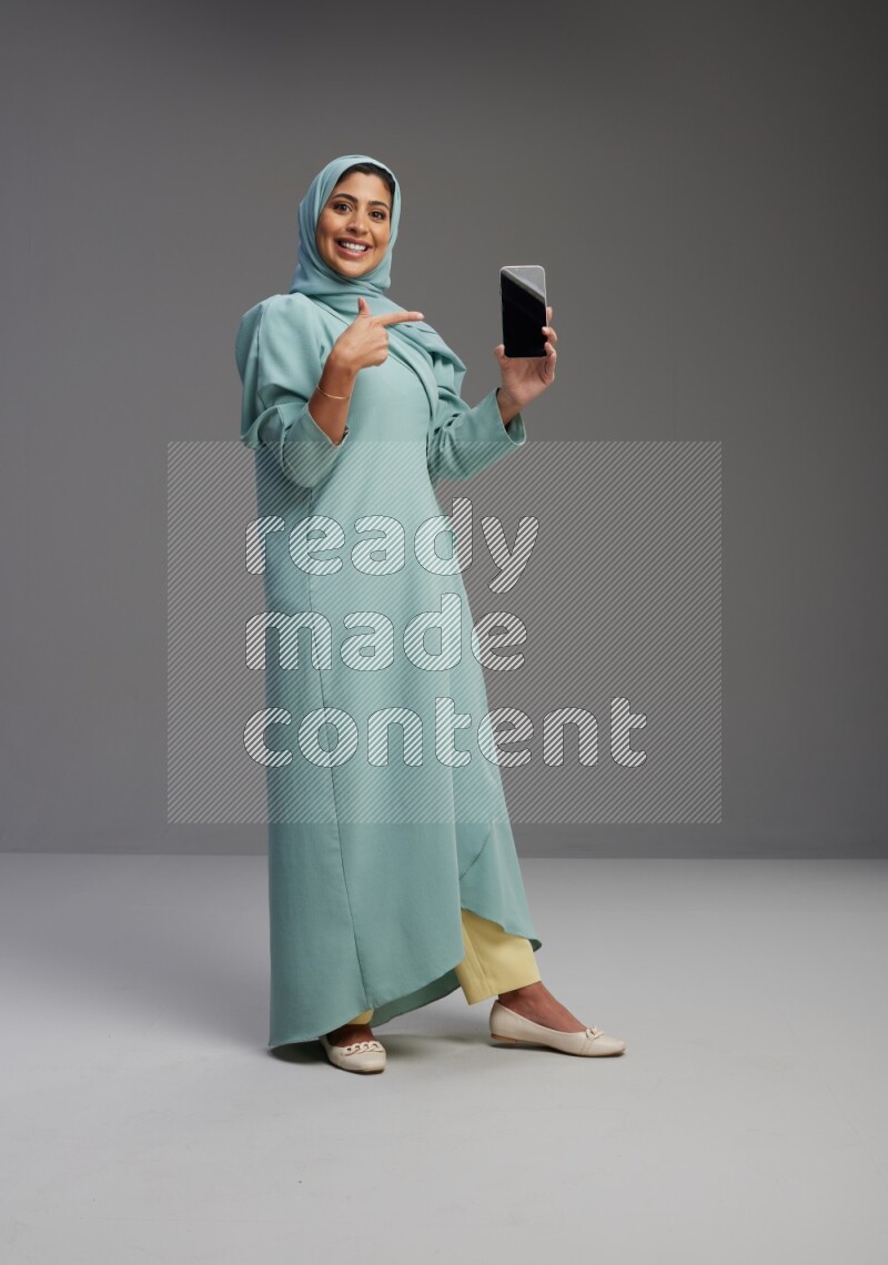 Saudi Woman wearing Abaya standing showing phone to camera on Gray background