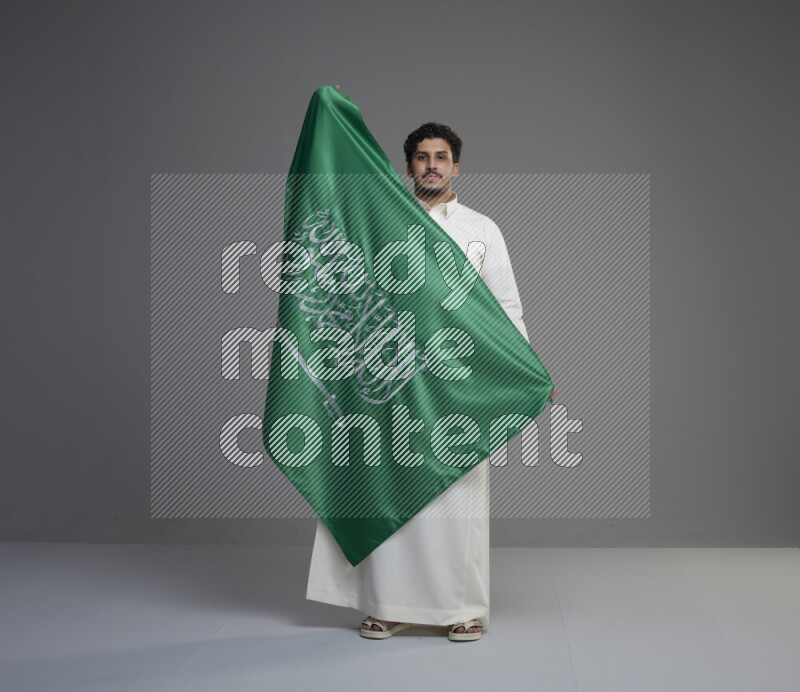A Saudi man standing wearing thob holding big Saudi flag on gray background