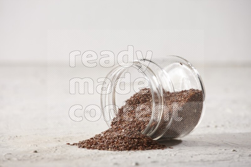A glass jar full of flax seeds flipped and seeds spread out on a textured white flooring