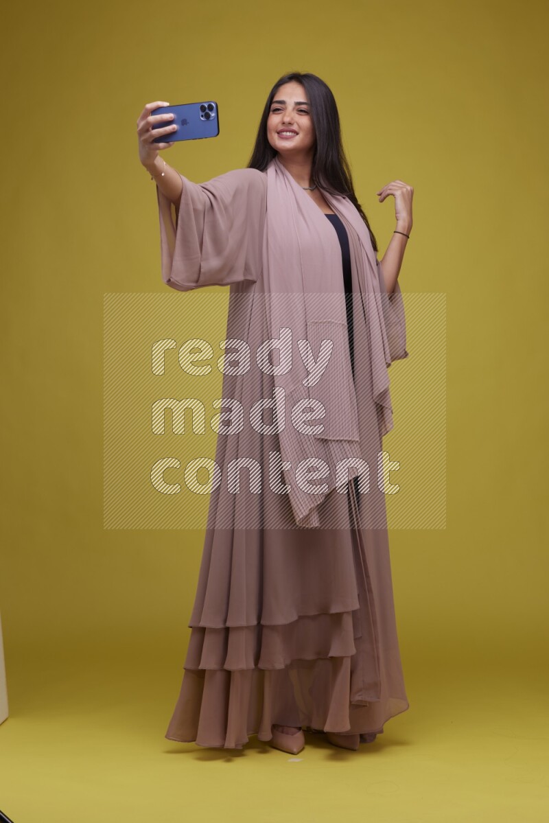A woman Taking a Selfie on a Yellow Background wearing Brown Abaya