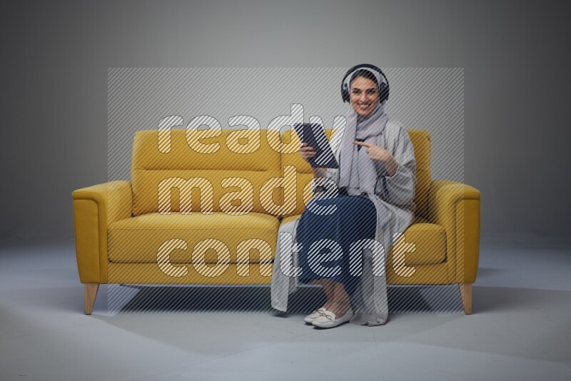 A Saudi woman wearing a light gray Abaya and head scarf sitting on a yellow sofa and using her phone while wearing headphones eye level on a grey background