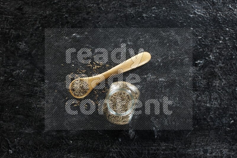 وعاء زجاجي للتوابل وملعقة خشبية ممتلئين بحبوب الكمون على خلفية سوداء