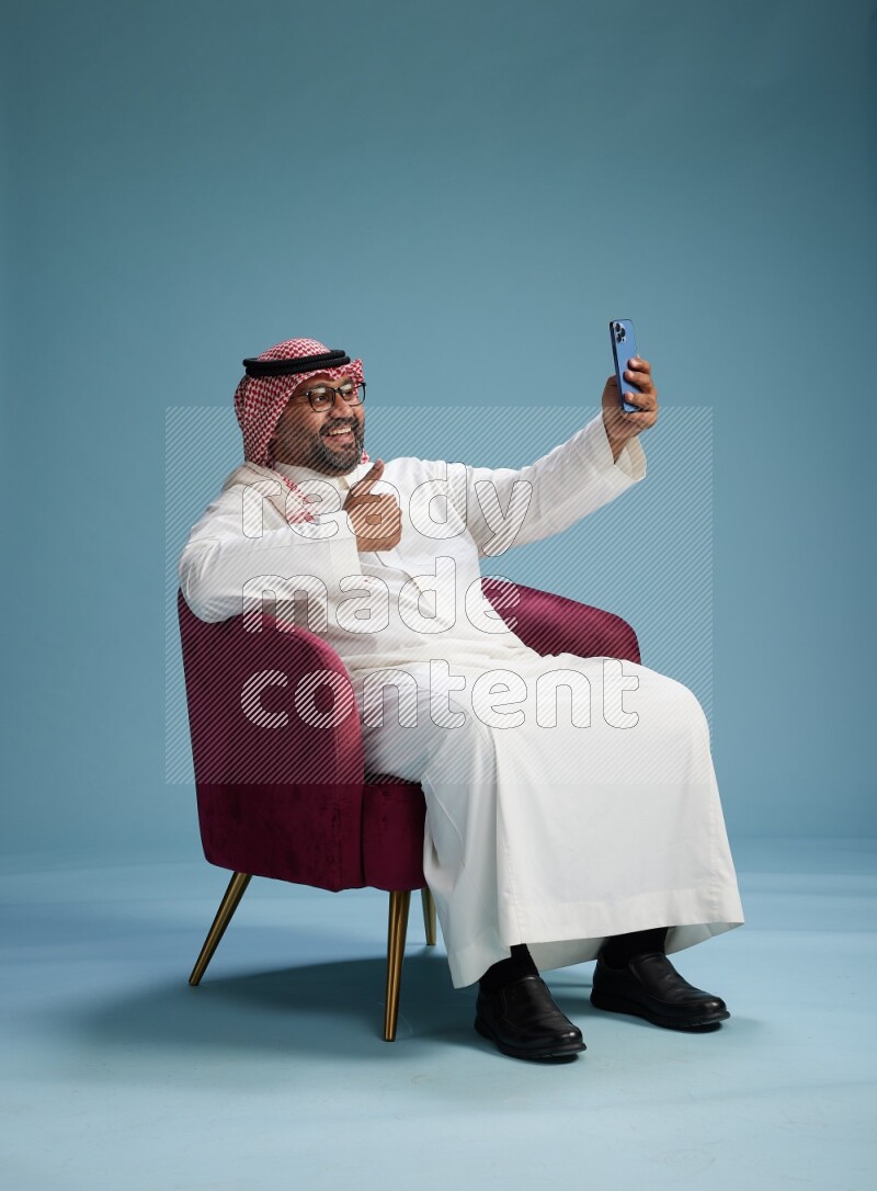 Saudi Man with shimag sitting on chair taking selfie on blue background
