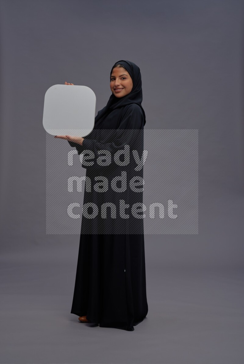 Saudi woman wearing Abaya standing holding social media sign on gray background