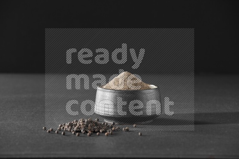 A black pottery bowl full of black pepper powder and black pepper beads on the floor on black flooring