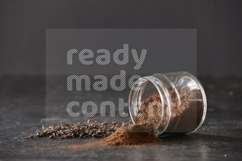 A flipped glass jar full of cloves powder with cloves spread on a textured black flooring