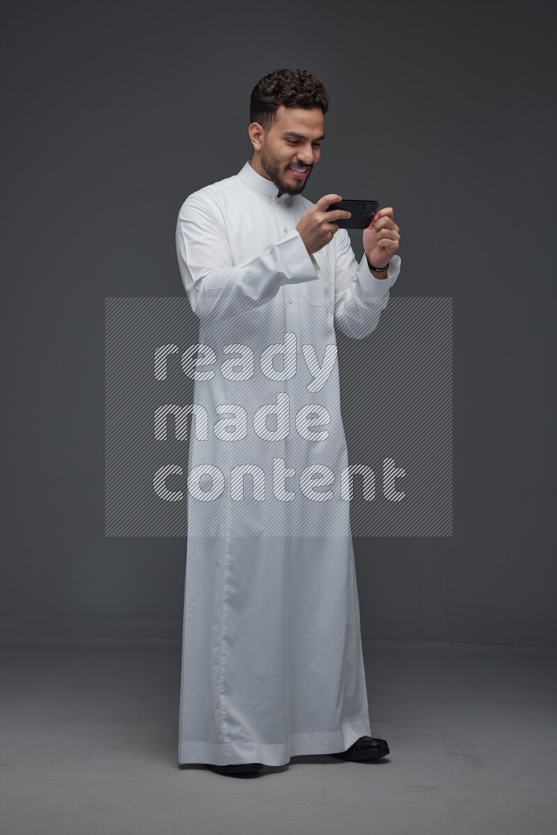 A Saudi man wearing Thobe and playing using his phone horizontally playing video games while standing and making different poses eye level on a gray background