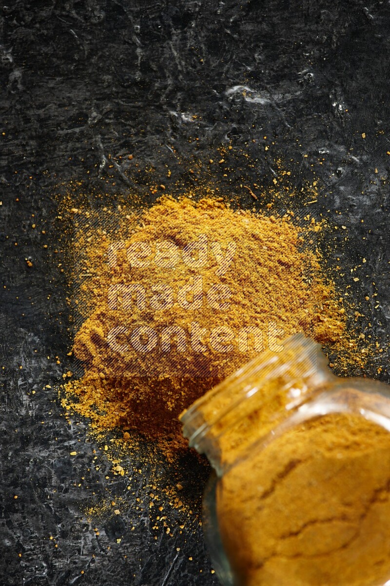 A flipped glass spice jar full of turmeric powder and powder spilled out of it on textured black flooring