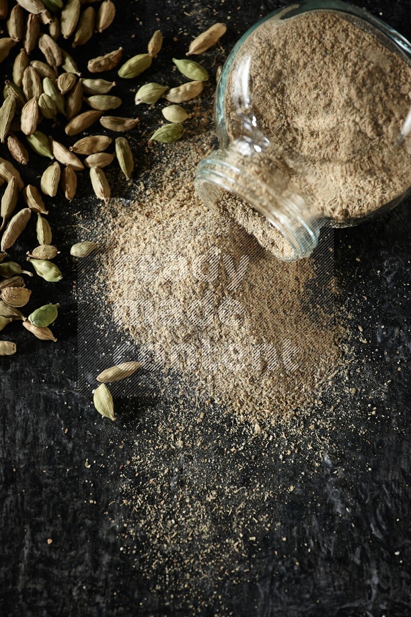 A flipped glass spice jar full of cardamom powder and powder spilled out of it with cardamom seeds spreaded on textured black flooring